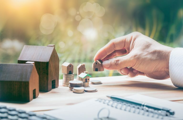 Man holding a model house, with other model houses and piles of coins on the desk, working out what deposit required