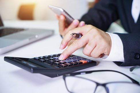 Business man at desk, using a calculator with pen in one hand and phone in other, getting organised for the Autumn