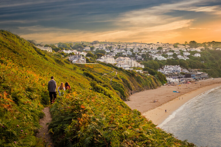 View of people walking in hilly seaside towards resort village in the bay at sunset; swimming beach and sunset sky in background